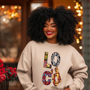 Puede incluir: Una sudadera beige con la palabra "Loc'd" impresa en la parte delantera en letras con patrones coloridos. Las letras est&aacute;n formadas por diferentes patrones geom&eacute;tricos e estampados de animales, incluidas rayas de cebra.