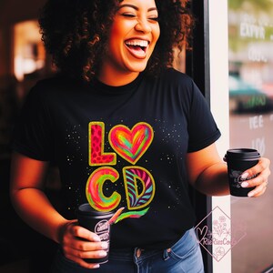 Könnte beinhalten: Schwarzes T-Shirt mit dem Text "LO CD" in bunten, lebendigen Buchstaben. Das "L" hat einen Leopardenmusterdruck, das Herz ist regenbogenfarben und das "CD" ist mehrfarbig. Eine Person trägt das Shirt und hält eine schwarze Kaffeetasse.