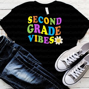 Puede incluir: Una camiseta negra con el texto "SECOND GRADE VIBES" en letras de colores y una margarita amarilla con una cara sonriente.