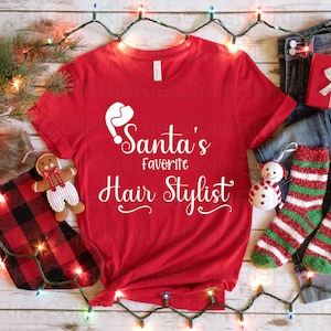 May include: Red t-shirt with white text that says "Santa's Favorite Hair Stylist". The shirt is surrounded by Christmas decorations, including a stocking, a snowman, and a gingerbread man.