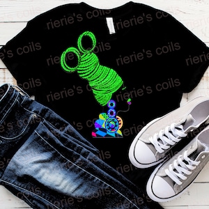 May include: Black t-shirt with a green and colorful graphic of a woman with a large afro hairstyle. The graphic is stylized and abstract. The t-shirt is on a white wooden surface with a pair of gray sneakers and blue jeans.
