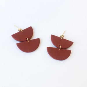 May include: A pair of terracotta-colored, half-circle earrings. Each earring has two stacked, semi-circular pieces connected by a gold-colored wire. The earrings have gold-colored posts.