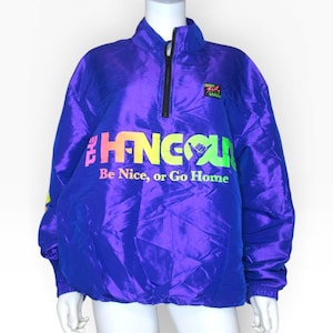 May include: A vibrant purple windbreaker jacket featuring a quarter-zip closure and a stand-up collar. The front displays the text THE HANGOUT in a multi-colored gradient font, with the phrase Be Nice, or Go Home printed in smaller letters below.