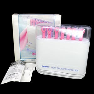May include: A white Conair Hot Sticks Traveller hair styling set with a clear plastic lid, featuring pink and purple heating rods. The set includes a white cardboard box with product imagery and a white paper instruction manual with black text.
