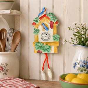 May include: A ceramic wall clock shaped like a house with a yellow frame, green leaves, and red grapes. Two blue birds are painted on the front. The clock face has black numbers and hands. Two white pinecone-shaped weights hang from red strings.