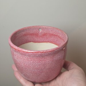 May include: A small, round ceramic bowl with a speckled pink exterior glaze and a smooth, off-white interior. The rim features two small, evenly spaced circular holes, suggesting it is designed for hanging as a wall-mounted planter or vessel.