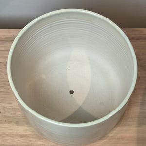 May include: A light grey ceramic planter with a round shape and a drainage hole at the bottom. The interior has horizontal ridges. The planter is suitable for indoor or outdoor use. The planter is sitting on a wooden surface.