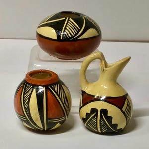 May include: Three small, hand-painted ceramic vessels. The pieces include a round vase, a small pitcher, and a spherical pot. Each is decorated with geometric patterns in black, cream, and orange against a brown or cream background. The pitcher has a handle and spout.