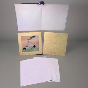 May include: An open notebook with a metal clip, a framed artwork with cranes, a gold-colored card, and three white envelopes. The artwork features two white cranes with black accents against a backdrop of orange, green, and black.