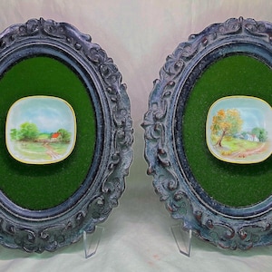May include: Two oval, ornate, dark gray frames with green backing, each holding a small, square, hand-painted landscape scene. The paintings depict a house, trees, and a path. The frames are displayed on clear stands.