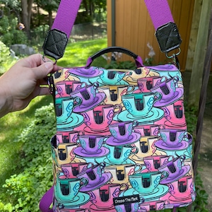 Puede incluir: Mochila morada y negra con un estampado colorido de tazas de té. La mochila tiene una cremallera y un pequeño bolsillo en la parte delantera. El texto "Dress The Park" está impreso en la parte delantera de la mochila.