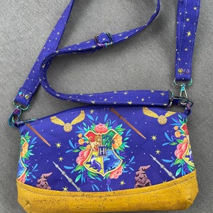 May include: A purple crossbody bag with a gold cork bottom and a matching adjustable strap. The bag features a repeating pattern of stars and magical elements, including a crest, wands, and a sorting hat. The bag has silver-tone hardware.