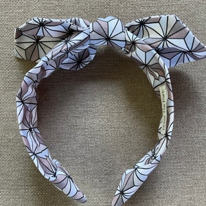 May include: A white headband with a geometric pattern in shades of gray and black. The headband is tied in a bow at the top and has the text "Dress The Part" printed on the side.