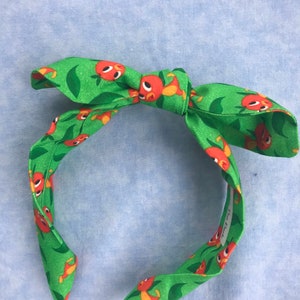 May include: Green headband with a red and orange cartoon bird print. The headband is tied in a bow at the top.