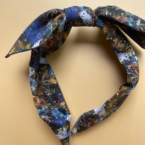 Puede incluir: Una diadema de tela con lazo, con un fondo azul oscuro con un estampado de búhos blancos, flores y estrellas. La diadema está diseñada con un nudo en la parte superior y dos extremos largos y puntiagudos.