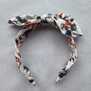 以下が含まれることがあります： オレンジ、ブルー、ブラックのカラフルなパターンが施された白いヘッドバンド。ヘッドバンドは、上部にリボンで結ばれています。