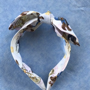 Puede incluir: Diadema de tela blanca con lazo, con un estampado repetido de osos marrones, detalles florales amarillos y figuras azules. El texto "Dress The Dark" está impreso en la diadema. La diadema está sobre una superficie azul claro.