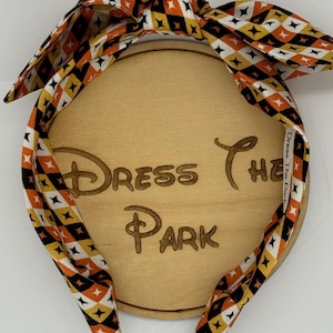 May include: A fabric headband featuring a repeating diamond pattern in orange, yellow, black, and white with small star accents. The headband is tied in a knot at the top and rests on a circular wooden plaque engraved with the text Dress The Park.