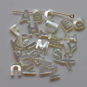 May include: A collection of white mother of pearl letter beads, each letter of the alphabet is represented. The beads are scattered on a white surface.