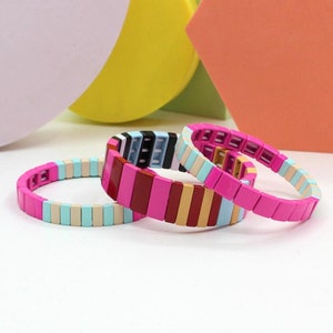 Op de afbeelding: Drie kleurrijke armbanden gemaakt van gestapelde plastic tegels. De armbanden zijn in tinten roze, blauw, beige, zwart en bruin.