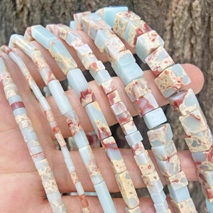 Puede incluir: Una colección de cuentas de jaspe de color azul claro y marrón con forma cuadrada. Las cuentas tienen un patrón moteado de marrón claro, blanco y rojo.