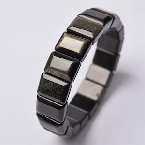 Könnte beinhalten: Ein schwarzes Obsidian-Armband mit rechteckigen, polierten Perlen. Die Perlen haben einen reflektierenden, metallischen Glanz mit Gold- und Silberakzenten. Das Armband wird auf weißem Hintergrund präsentiert.