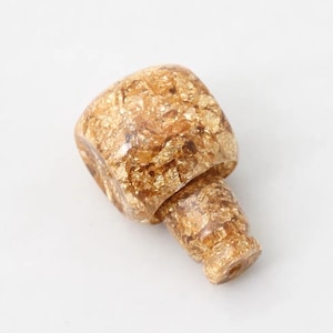 Peut inclure: Un bouchon de bouteille décoratif avec un motif de feuilles d'or. Le bouchon est en résine transparente avec des flocons de feuilles d'or incrustés. Il a un dessus carré et une base cylindrique.