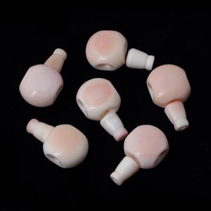 Peut inclure: Sept têtes de poupée roses et blanches avec une surface lisse et légèrement texturée. Les têtes sont toutes de formes et de tailles différentes, mais elles ont toutes un style similaire.