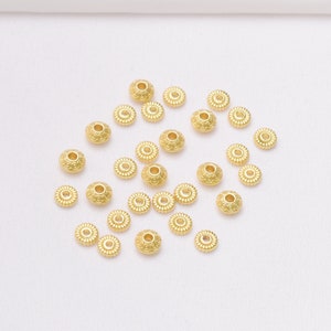 Pode incluir: Uma coleção de contas de metal douradas com um design decorativo. As contas estão espalhadas sobre uma superfície branca.