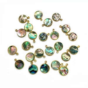 Könnte beinhalten: Eine Sammlung runder Abalone-Muschel-Anhänger mit goldfarbenen Metallrahmen und kleinen Schlaufen zum Befestigen an Schmuck. Die schillernden Muscheln zeigen eine Reihe von Farben, darunter Blau, Grün und Lila, wodurch ein schimmernder Effekt entsteht. Die Anhänger sind auf einer weißen Oberfläche verstreut.