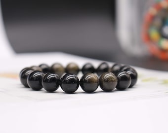 Könnte beinhalten: Ein schwarzes Perlenarmband mit einer Mischung aus glänzenden und strukturierten Perlen. Das Armband besteht aus runden, polierten Perlen, von denen einige einen goldenen Schimmer haben. Das Armband ist ein Schmuckstück.