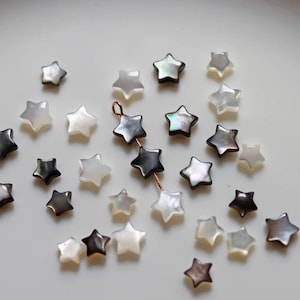May include: A collection of white and black star-shaped beads made of mother of pearl. The beads are scattered on a white surface.