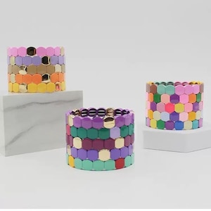 Op de afbeelding: Drie kleurrijke armbanden gemaakt van zeshoekige kralen. De armbanden zijn gestapeld op witte platforms. De armbanden zijn gemaakt van verschillende kleuren, waaronder paars, groen, geel, oranje, roze en blauw.