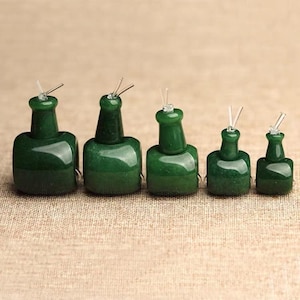 Puede incluir: Cinco adornos en forma de botella de jade verde oscuro de varios tamaños. Cada uno tiene un pequeño tapón transparente, similar al vidrio, con dos finas varillas transparentes, similares al vidrio, que se extienden hacia arriba. Las botellas están dispuestas en fila.