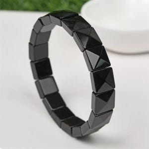 Può includere: Un braccialetto nero composto da perline quadrate a forma di piramide. Le perline sono infilate insieme per formare una fascia flessibile. Il braccialetto è esposto su una superficie bianca, con uno sfondo sfocato.