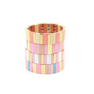Può includere: Pila di braccialetti colorati con accenti dorati. Ogni braccialetto è fatto di piastrelle rettangolari rosa, bianche e beige.