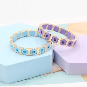 May include: Two gold bracelets with flower-shaped links. One bracelet has light blue enamel and turquoise gemstones, the other has purple enamel and clear gemstones.