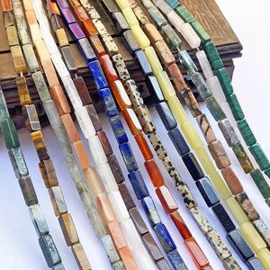 May include: Assortment of 16 strands of gemstone beads, each strand featuring a different color and pattern of rectangular beads. The beads are arranged in a row, showcasing the variety of colors and textures.