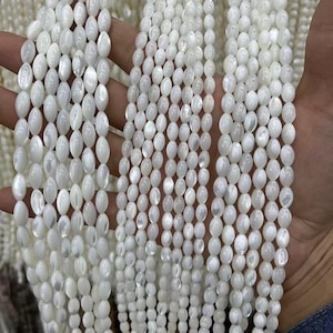 May include: Strands of lustrous, white oval-shaped beads. The beads are strung together, creating a flowing, textured appearance. The beads are held in a hand, showcasing their size and shape. The overall theme is jewelry making or beadwork.