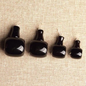 Peut inclure: Quatre petits pendentifs noirs brillants en forme de bouteille de tailles variables sont disposés en rangée. Chaque pendentif a un corps arrondi et un col fin, avec une fine ficelle transparente attachée en haut. Les pendentifs sont présentés sur une surface texturée de ton neutre.