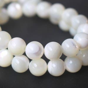 Puede incluir: Primer plano de un collar de cuentas redondas, blancas e iridiscentes. Las cuentas lisas y pulidas están ensartadas, formando una forma circular. Las cuentas, de color blanco cremoso, reflejan la luz, creando un brillo sutil. Cada cuenta mide unos 0,8 cm de diámetro.