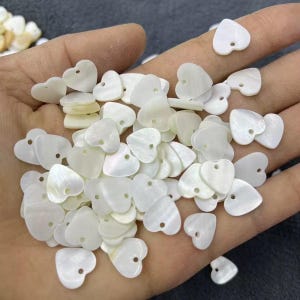 May include: A collection of small, heart-shaped, white shell beads with a hole at the top for stringing. The beads have a pearlescent sheen and are scattered across a hand.
