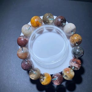May include: A bracelet made of spherical gemstones in various colours, including orange and brown, with internal inclusions. The beads are displayed on a white surface. The bracelet's design highlights the natural beauty of the stones, with each bead approximately 1.5 cm in diameter.