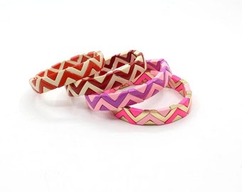 Puede incluir: Cuatro pulseras apiladas unas sobre otras con un patrón de chevron colorido. Las pulseras están hechas de azulejos de cerámica con un acento dorado. Las pulseras son en tonos de rosa, rojo y blanco.