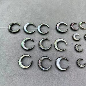Pode incluir: Uma coleção de contas de concha em forma de lua crescente, iridescentes em preto e branco. As contas são enfiadas em um fio fino e estão prontas para serem usadas na fabricação de joias.