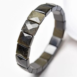 May include: A black and gold obsidian bracelet. The bracelet is made of square-cut beads with a polished finish. The beads have a reflective, metallic sheen, and the bracelet is set against a white background.