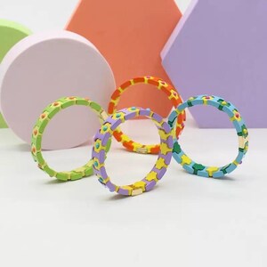 Op de afbeelding: Vier kleurrijke armbanden met een bloemenontwerp. De armbanden zijn gemaakt van keramische tegels en hebben een gouden sluiting. De armbanden zijn in een cirkel op een wit oppervlak gerangschikt.