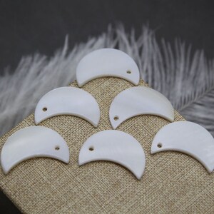 May include: Six white half-moon shaped shell charms with a hole in the top for stringing. The charms are arranged in a semi-circle on a beige fabric background.