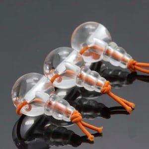 Peut inclure: Trois objets transparents en forme de bulbe, attachés à des cordons orange. Chaque objet a un sommet arrondi et une base striée. Les cordons sont noués à la base. Les objets sont disposés sur une surface réfléchissante.