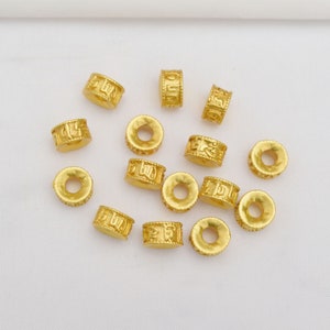 Könnte beinhalten: Eine Sammlung von 14 goldfarbenen Metallperlen mit eingravierten chinesischen Schriftzeichen. Die Perlen sind auf einer weißen Oberfläche verstreut.